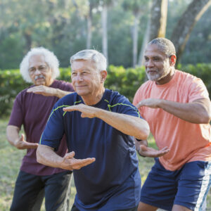 A group of three multi-ethnic senior men in the park practicing tai chi. The focus is on the caucasian man standing in the middle. He is in his 70s.