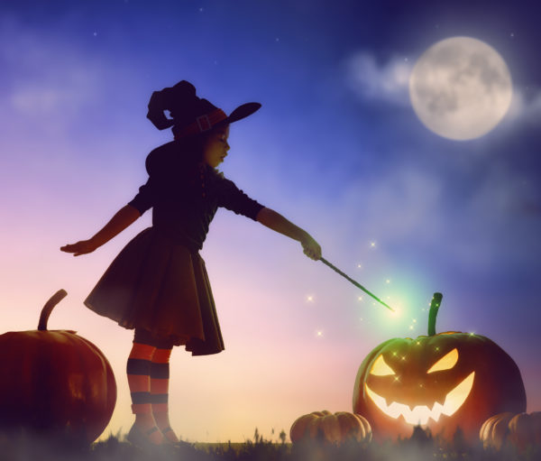 Girl in witch costume pointing magic wand at jack-o-lantern